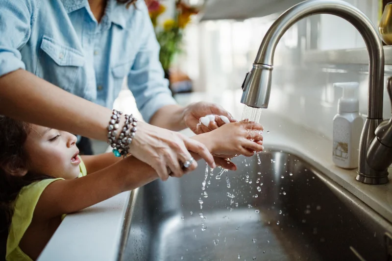 Woman helping a young girl wash her hands in a kitchen sink.